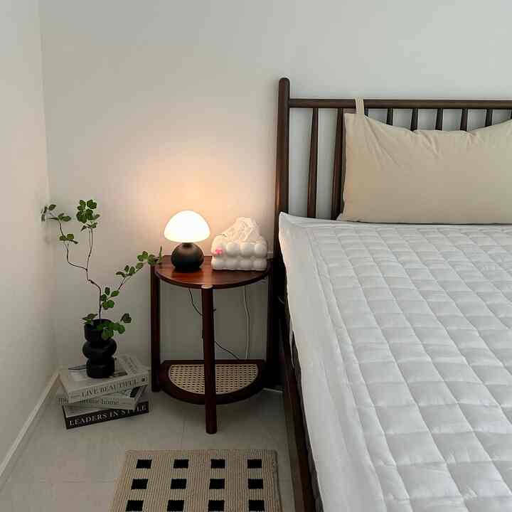 ホワイトと木目調のモダンな寝室、木製ベッドフレームとナイトテーブル、テーブルランプと植物が落ち着いた雰囲気を演出