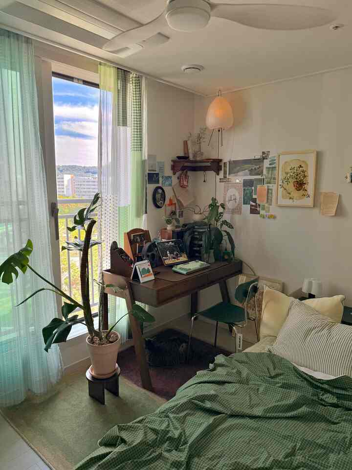 白い壁と緑のカーテンがある小さな寝室、木製のデスクと植物が配置されたナチュラルで居心地の良い空間