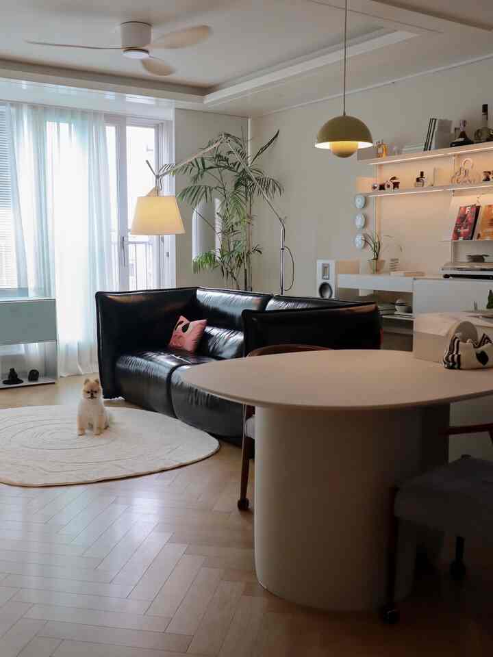 Natural modern living room with white walls and brown wood flooring, featuring black leather sectional sofa, round dining table, and a pet dog