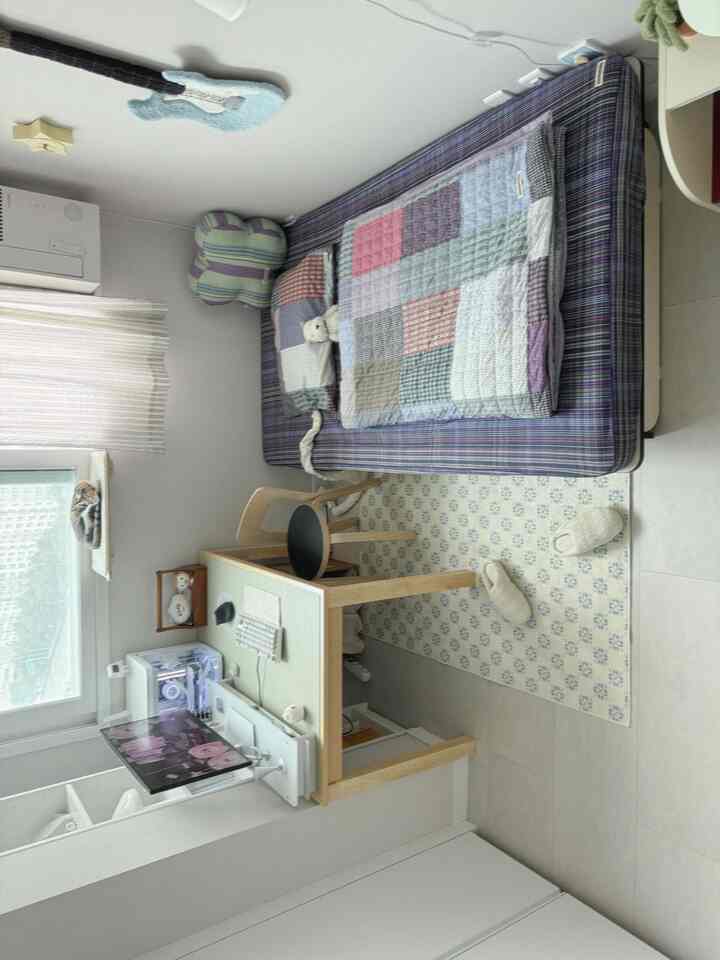 明るいナチュラルトーンのワンルーム寝室、木製デスクとコンピューター、チェック柄の寝具、窓際にキャットベッドとカーテンが配置された雰囲気のある空間