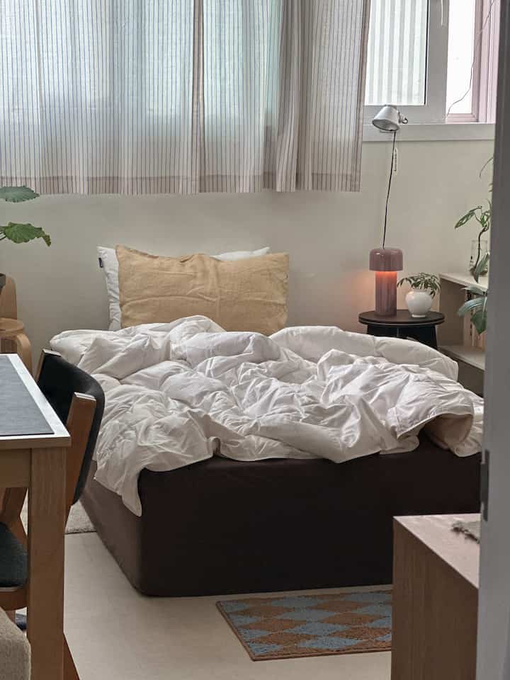 ホワイトとベージュのトーンのナチュラルモダンな寝室、中央に枕と布団のあるベッドとサイドテーブルのランプが配置された落ち着いた空間