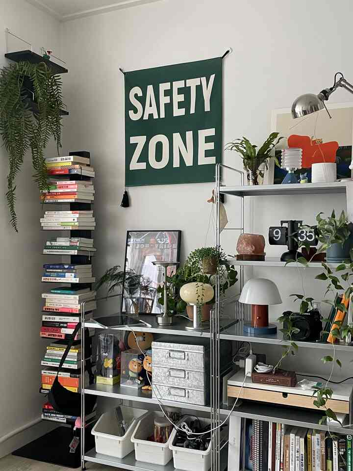 白い壁と緑のファブリックポスター、金属の棚に本や植物が配されたシンプルでミッドセンチュリーモダンなリビングコーナー