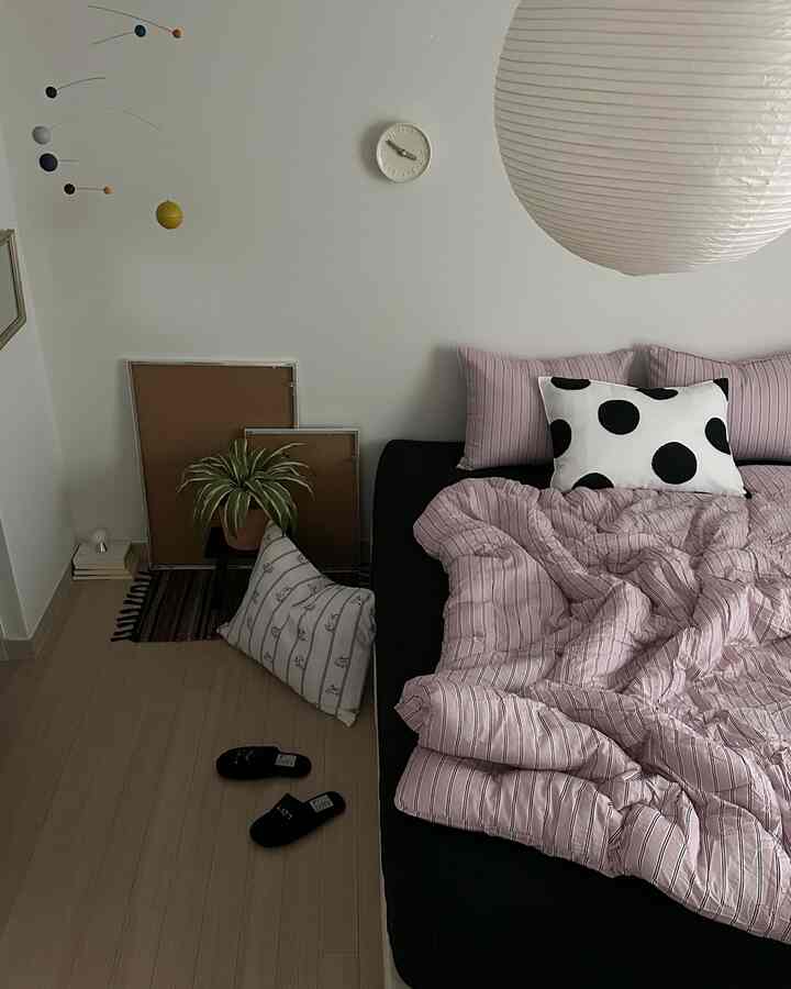 A cozy bedroom with white walls and light wood flooring, featuring a black sofa bed with pink striped bedding, ceiling mobile, and potted plant