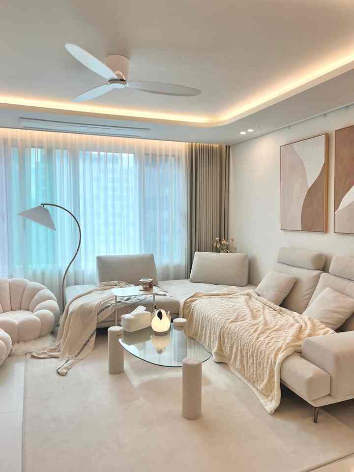 Soft beige-toned living room featuring a corner sofa, round stool, and glass coffee table with a modern, cozy atmosphere