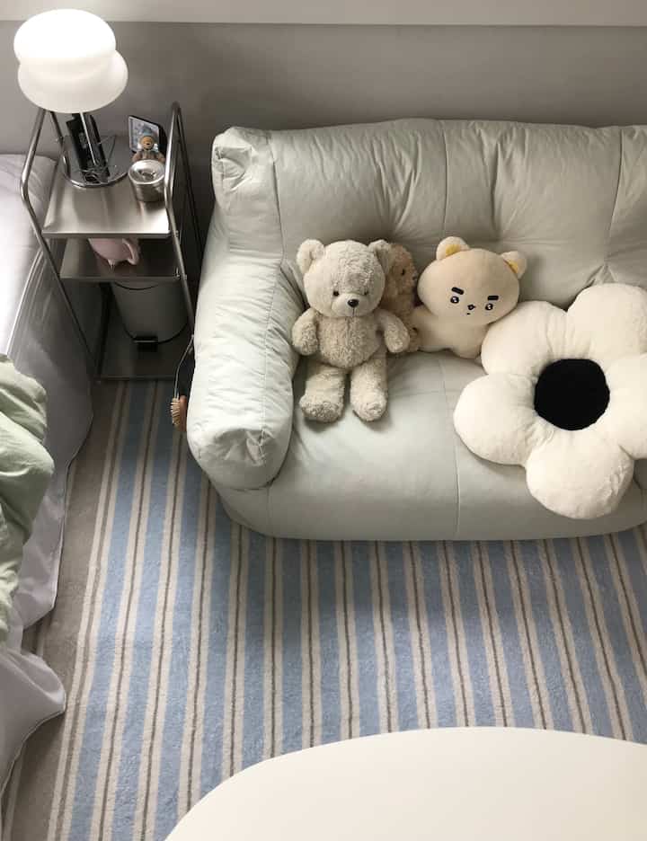 ベージュのビーズクッションソファと青いストライプのラグ、ステンレス製のサイドテーブルと柔らかなクッションが配置されたシンプルな寝室のインテリア