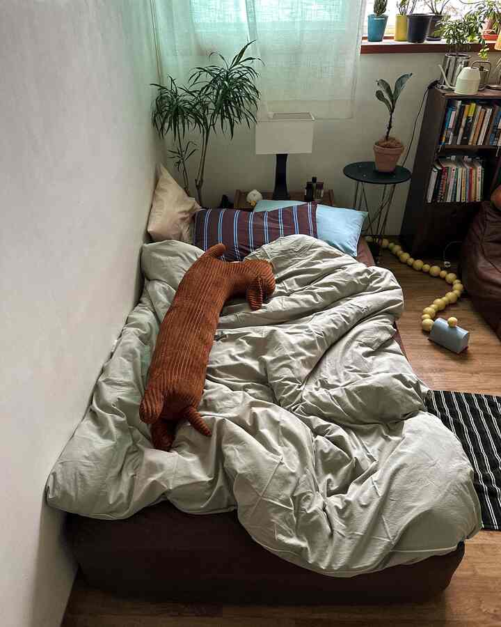 ナチュラルなウッドトーンと植物が調和した落ち着いた寝室、ベッドの上に犬のぬいぐるみと寝具が配置された印象的な空間