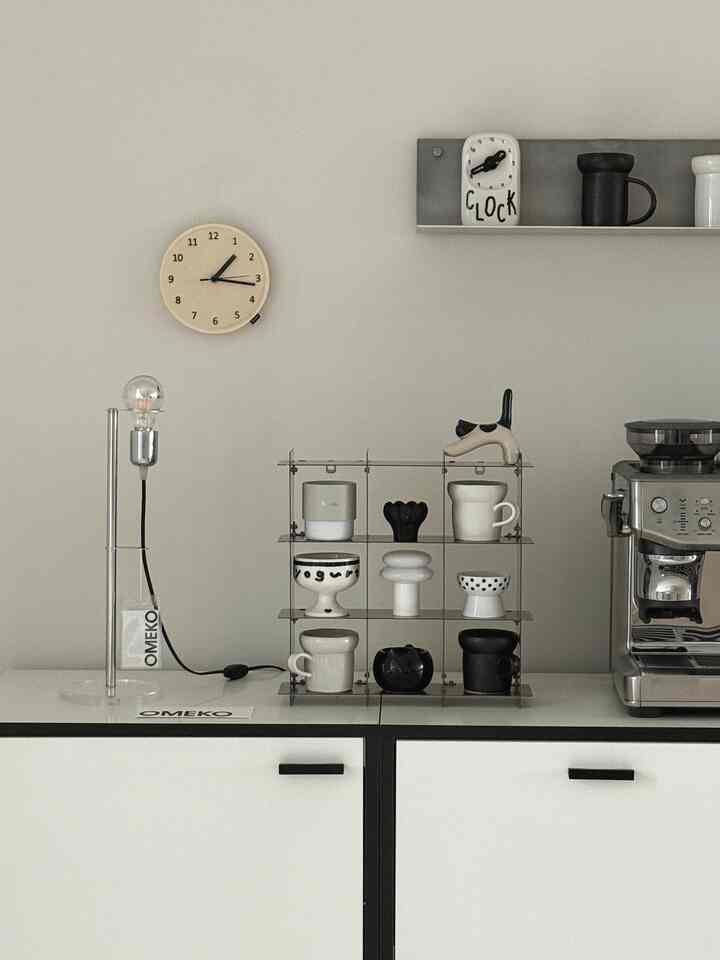 白い壁と白黒のキッチンキャビネットの上に、ユニークな形のテーブルランプとアルミ製の壁棚、コーヒーマシンが配置されたモダンなキッチンスペース