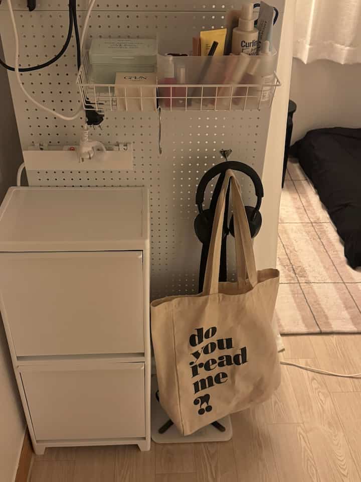 白とナチュラルカラーのワンルーム、化粧品収納用のパーティション、小さな収納キャビネット、バッグとヘッドホンが配置された雰囲気のある寝室