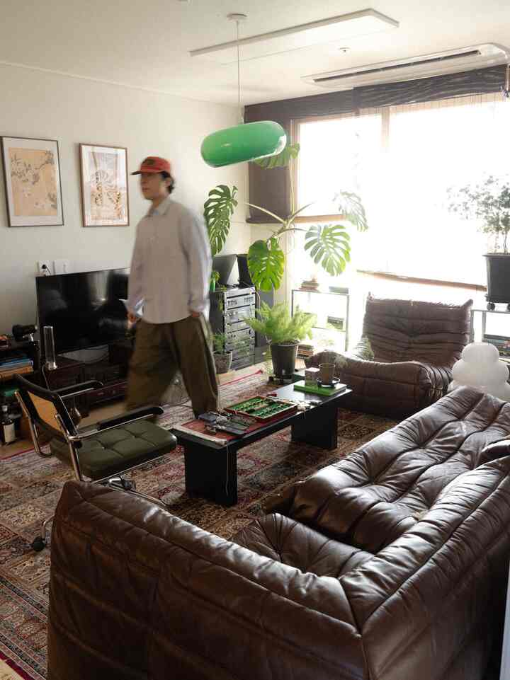 ブラウンレザーのソファとアームチェアが配置されたリビングルーム、大きなグリーンのペンダントライトと植物が印象的なヴィンテージ風インテリア