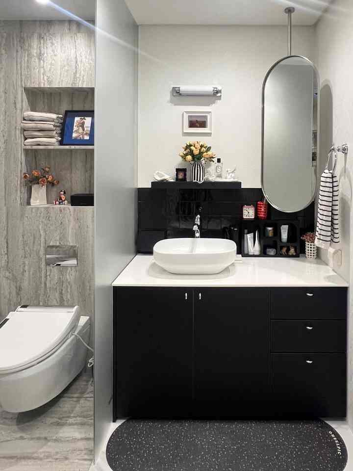 A clean bathroom in white and black tones featuring a central washbasin with an oval mirror, wall shelves, and a bath mat on the floor