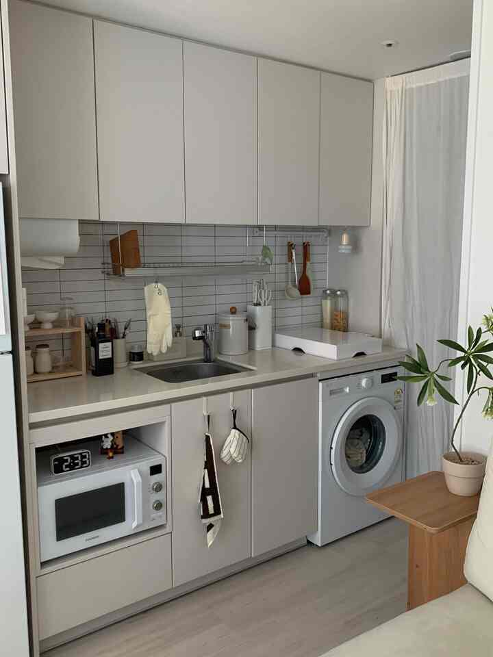 ホワイトとナチュラルトーンのワンルームキッチン、整理された収納と洗濯機が配置された雰囲気のある空間
