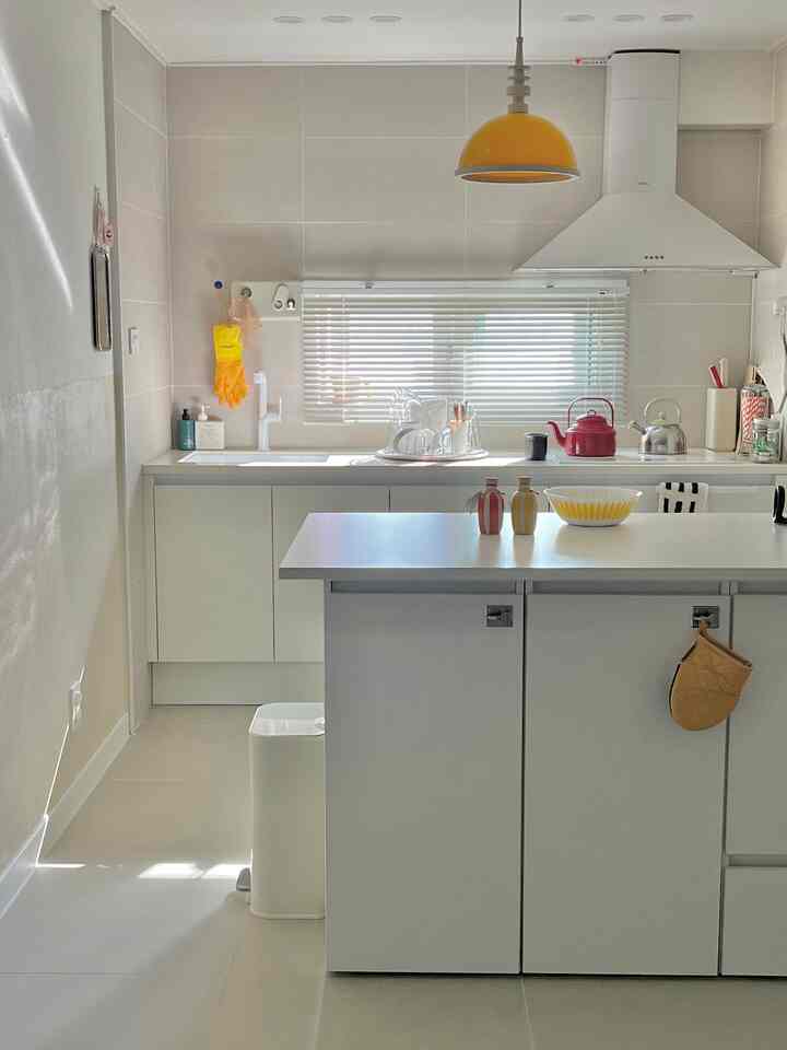 白とナチュラルトーンのキッチン、中央にダイニングテーブルとブラインドが配置された清潔でミニマルな雰囲気のインテリア