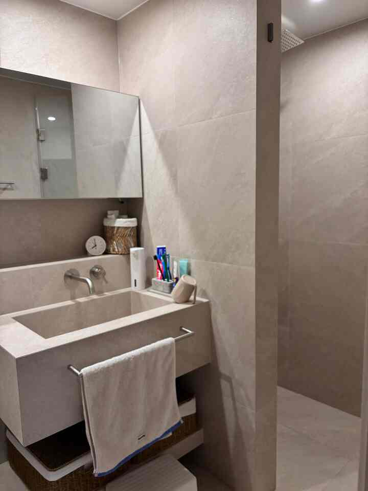 ベージュの色調のバスルーム、シンプルな洗面台とタオル、時計、収納バスケットが整然と配置されたホテルライクなインテリア