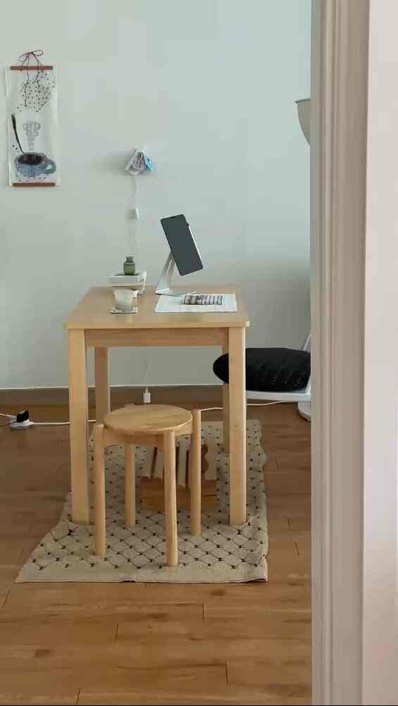 ナチュラルな木目調の床と壁に、木製のデスクと丸いスツール、黒のクッションチェアが配置された一人暮らしのホームオフィス空間