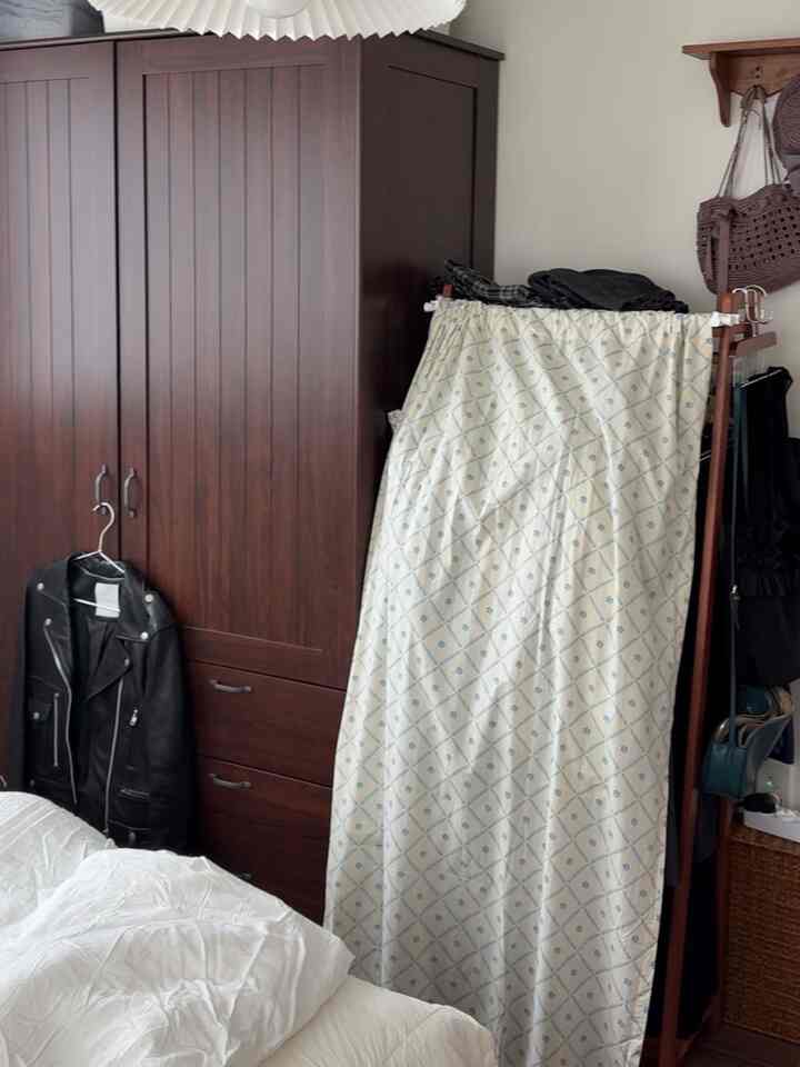 白と茶色を基調とした寝室、収納アイデアが感じられるワードローブとカーテン付きのハンガーラックが配置された落ち着いた空間