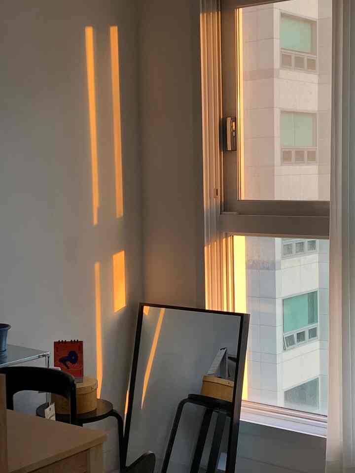 温かいオレンジ色の陽光が差し込むホワイトの小さな部屋、黒いスツールと鏡が配置されたコンパクトな空間