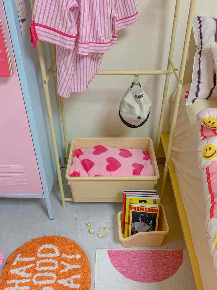 ピンクとベージュのトーンで統一された寝室の一角に、子供用の服や寝具、ハンガーラック、収納ボックスが配置された雰囲気のある室内