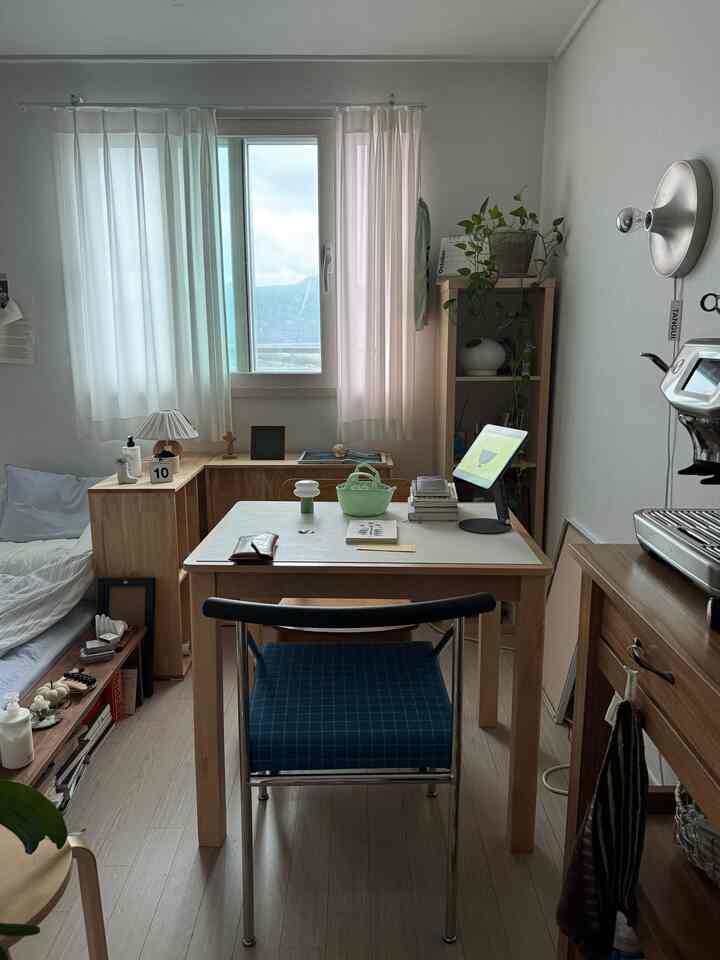 明るい木製家具とベージュのカーテンがある一人暮らし用の寝室兼作業スペース