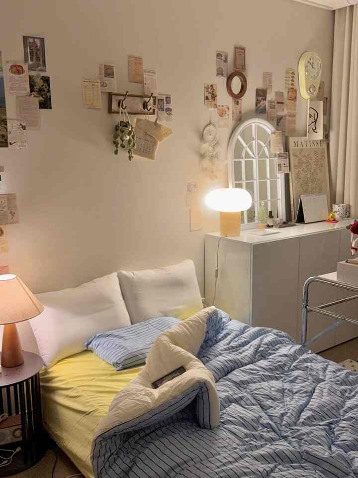 Cozy bedroom with soft white and yellow tones, featuring a nightstand table lamp and vintage-style posters decorating the wall