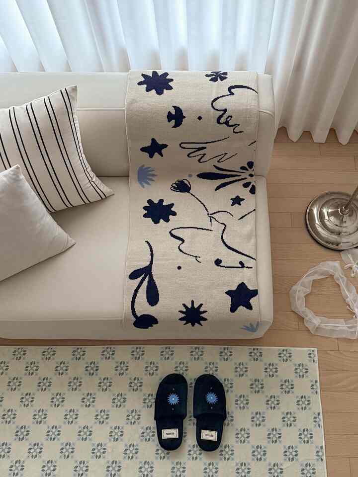 Beige and black-toned living room featuring a two-seat sofa with patterned throw and cushions, patterned rug and navy slippers on wood floor creating a cozy atmosphere