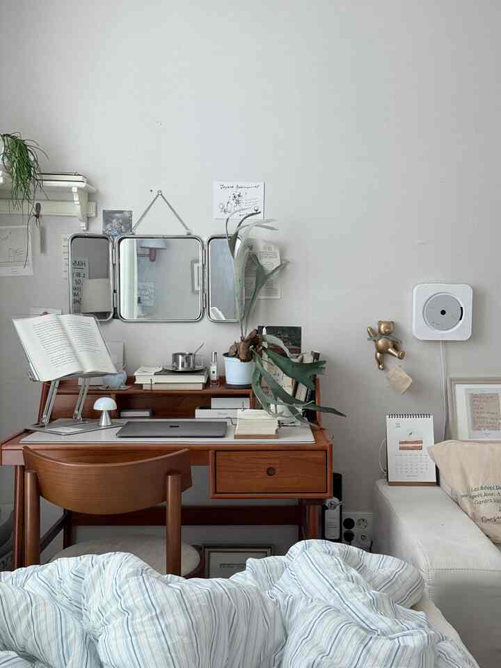 白い壁と木目調のデスクが配されたナチュラルカラーのホームオフィス空間、ヴィンテージ風の鏡とプランテリアが調和した居心地の良い書斎スペース