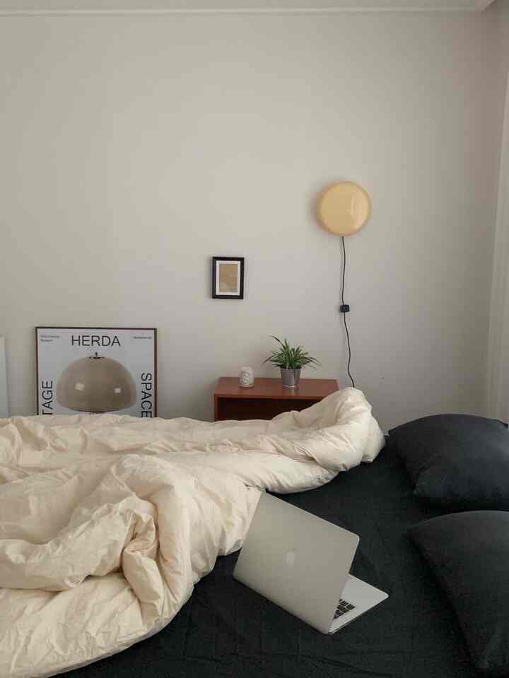 白と黒のトーンが特徴の寝室で、中央にクリーム色の掛け布団が敷かれたベッドがあり、壁にミッドセンチュリーモダンのポスターと黄色い照明が配置された印象的な空間