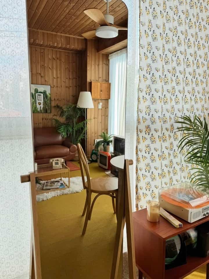 暖かみのあるウッドトーンとイエローのカーペットが特徴のワンルームリビング、ヴィンテージ家具と植物が配置された印象的な空間