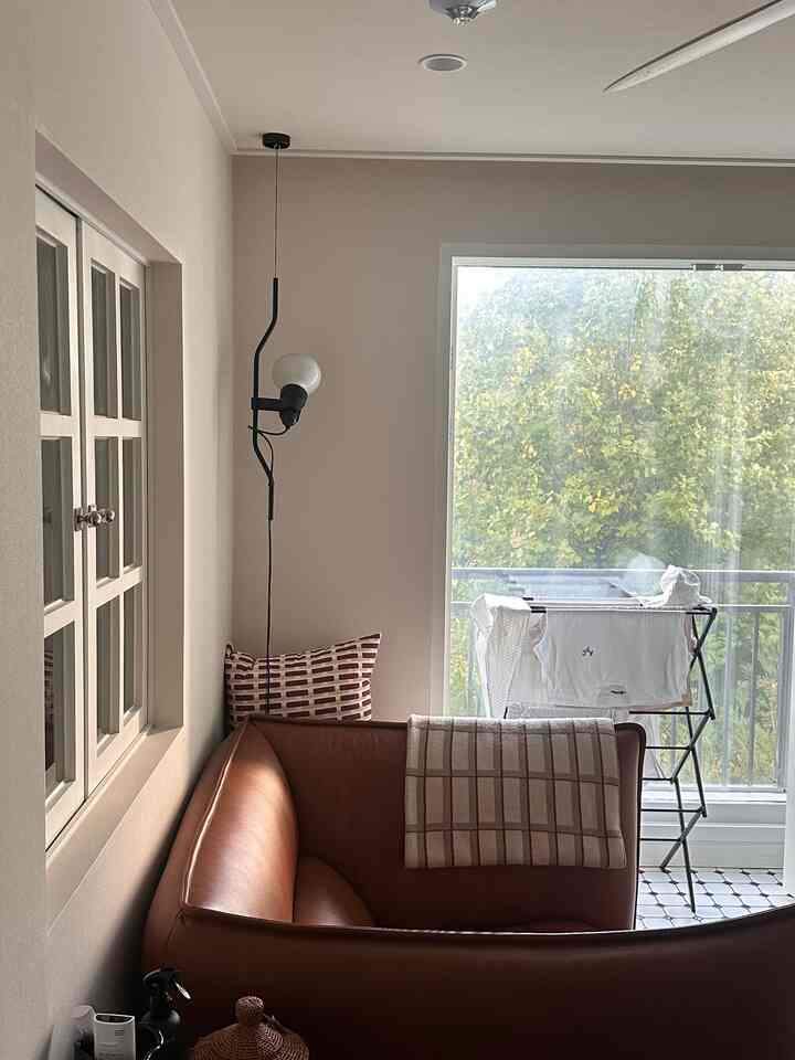 ブラウンレザーのソファと壁掛け照明がある明るくナチュラルなリビング、バルコニーの向こうに緑の木々が見える