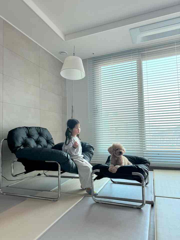 ホワイトの壁とブラインドがあるミッドセンチュリーモダンのリビング、黒いクッションのラウンジチェアとオットマンに子供と犬が座っている印象的な空間