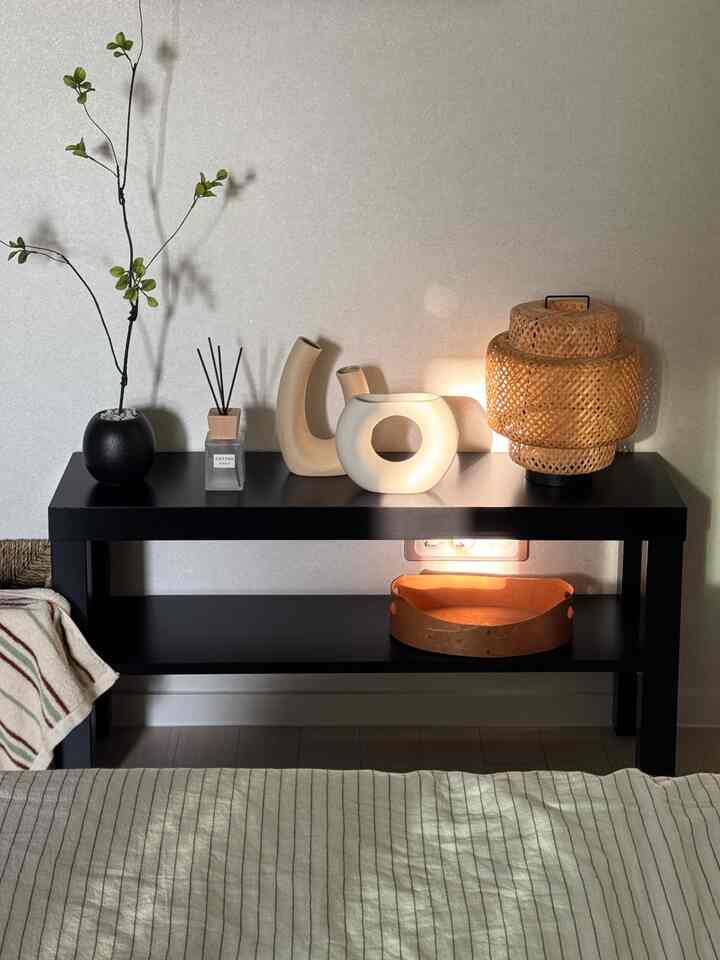 黒いラックの上に植木鉢、ディフューザー、陶器のオブジェ、ランプが置かれた落ち着いた寝室空間