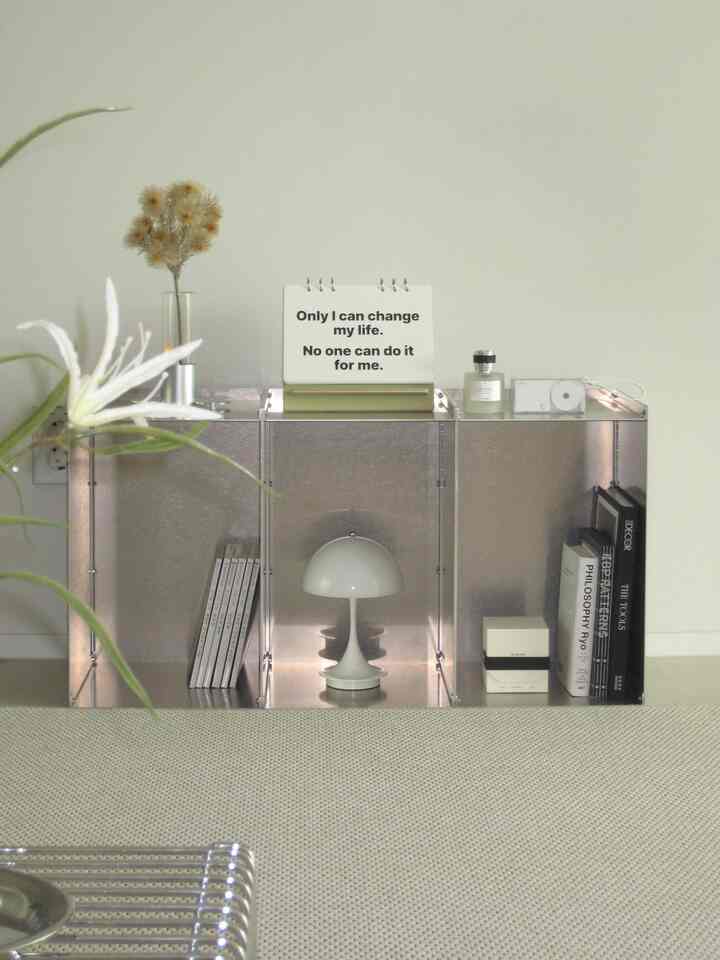 ホワイトとシルバーのトーンのシンプルな本棚が並び、テーブルランプと本、整然とした小物が配置された印象的な空間