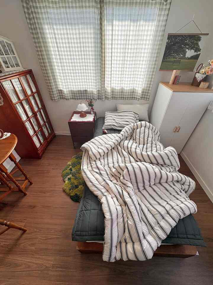 ベージュ色の壁と木材の床の寝室、ディープグリーンの寝具とストライプ模様の布団が置かれた温かみのある空間
