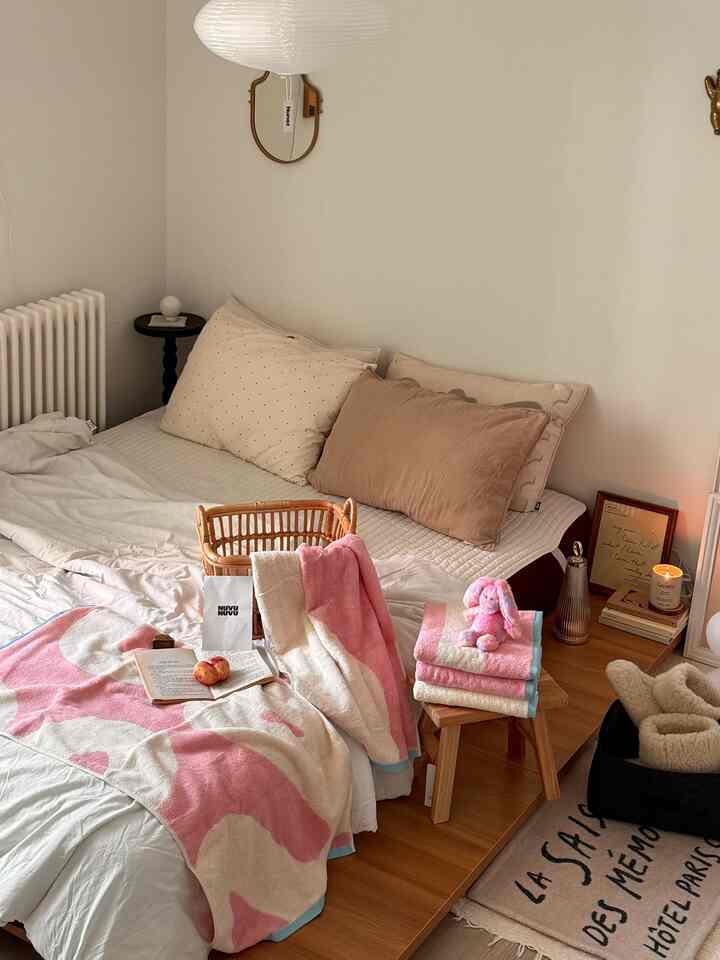 アイボリーとピンクのトーンが調和した寝室、ヘッドボードなしのベッドと温かみのあるタオル、穏やかな照明が配置された雰囲気のあるインテリア