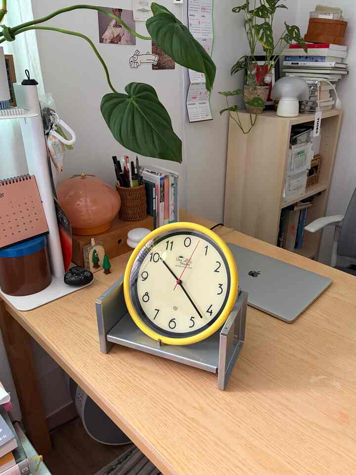 白い壁と木目調のデスクが配置されたコンパクトでモダンなホームオフィス空間、黄色い時計と植物がアクセントとして置かれている