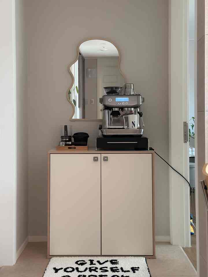 ベージュとナチュラルウッドトーンのキッチンスペース、ミッドセンチュリーモダンスタイルの波型ミラーとエスプレッソマシンが配置された清潔感のあるおうちカフェ