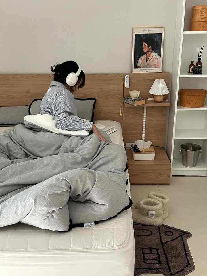 グレーの寝具とナチュラルな木目調家具が調和したシンプルモダンな寝室、ベッドで読書をする人がいる印象的な空間