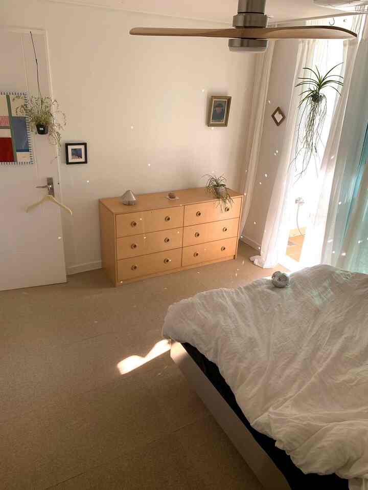 ホワイトとウッドトーンが調和する寝室、中央のチェストとベッド、植物が配置された自然で落ち着いた雰囲気の空間