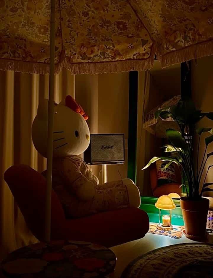 暖かい黄色の照明とヴィンテージ風フロアランプがある居心地の良いリビング、大きなハローキティのぬいぐるみと観葉植物が配置された空間