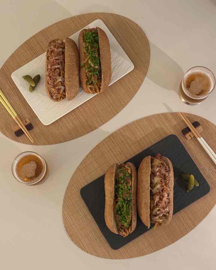 ブラウンとホワイトのトーンのダイニングテーブルに、パンに包まれた日本風焼きそばとビールグラスが配置された、雰囲気のあるホームバー風の食事空間