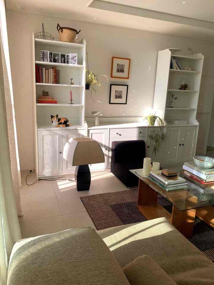 暖かい自然光が差し込む白と茶色の色調のリビング、ソファとガラステーブル、本棚と額縁が配置された雰囲気のある空間