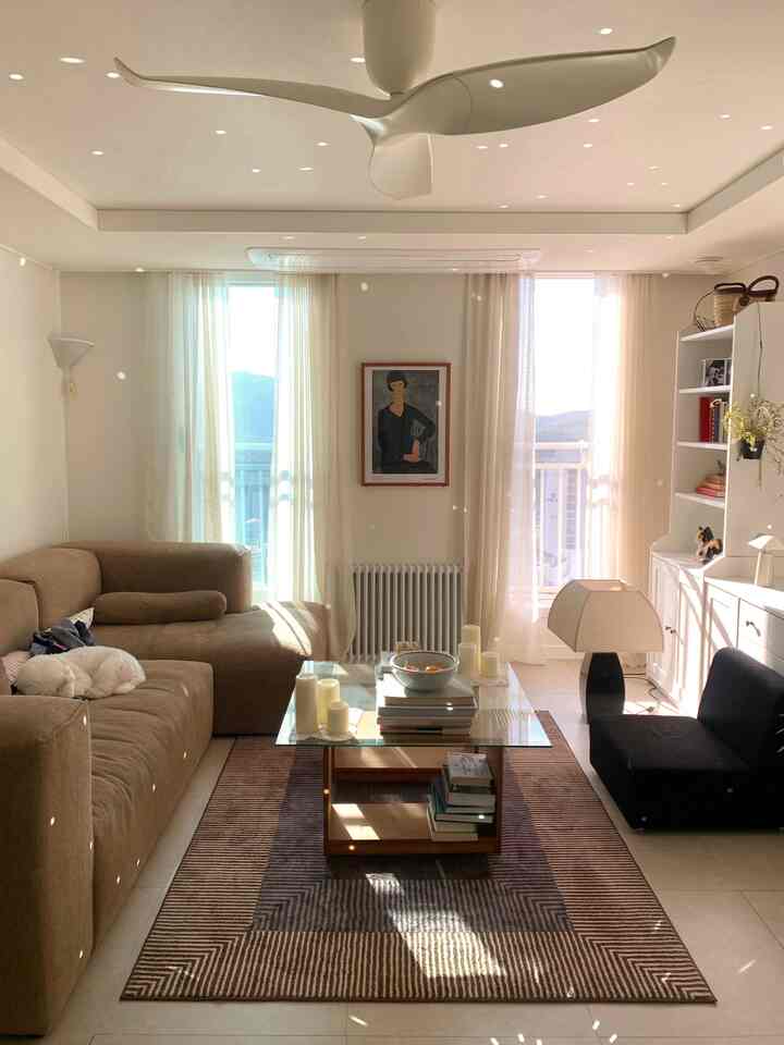 柔らかいブラウンとホワイトの色調のリビング、広いソファとリネンカーテン、中央にガラスのコーヒーテーブルとヴィンテージ風の額が配置された温かみのある空間