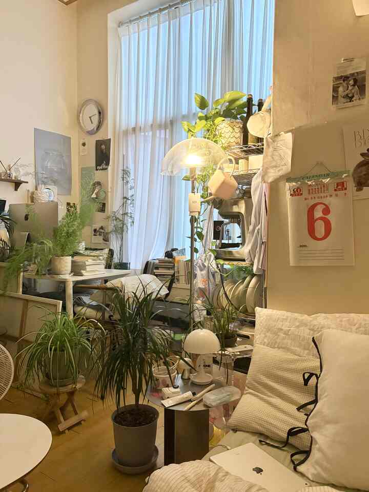 白い壁と自然の色調が調和する小さなリビング、ソファとデスク、多様な植物が配置された雰囲気のある空間