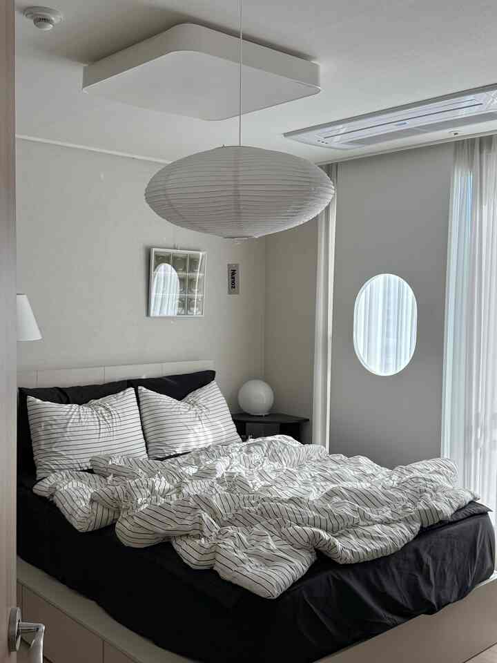 ナチュラルカラーの6畳寝室、黒いベッドとストライプ柄の寝具、白いペンダントライトとカーテンが配置されたモダンなインテリア