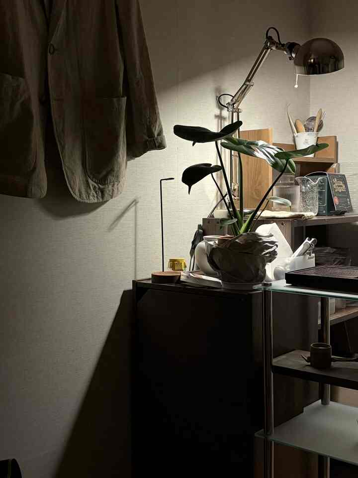ナチュラルカラーとブラウン調の落ち着いたコーナー収納スペース、大きな植物とシルバーの作業灯が配置されたミニマルな雰囲気の部屋