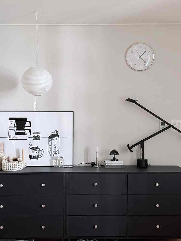 白い壁と黒いチェストが配置されたモダンな寝室、時計とテーブルランプが調和する雰囲気のあるインテリア