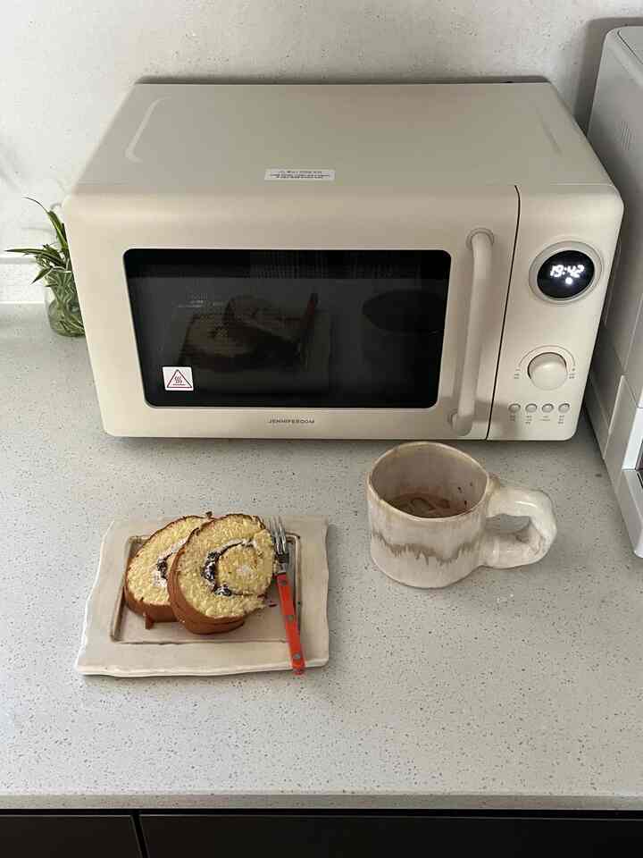 ベージュの電子レンジと白い調理台の上に皿、フォーク、マグカップが置かれたシンプルなキッチンの風景