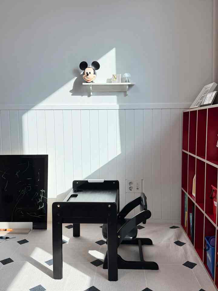 ホワイトとブラックのトーンの子供部屋、中央に黒い子供用机と椅子、右側に赤い本棚が配置されたモダンな勉強用インテリア