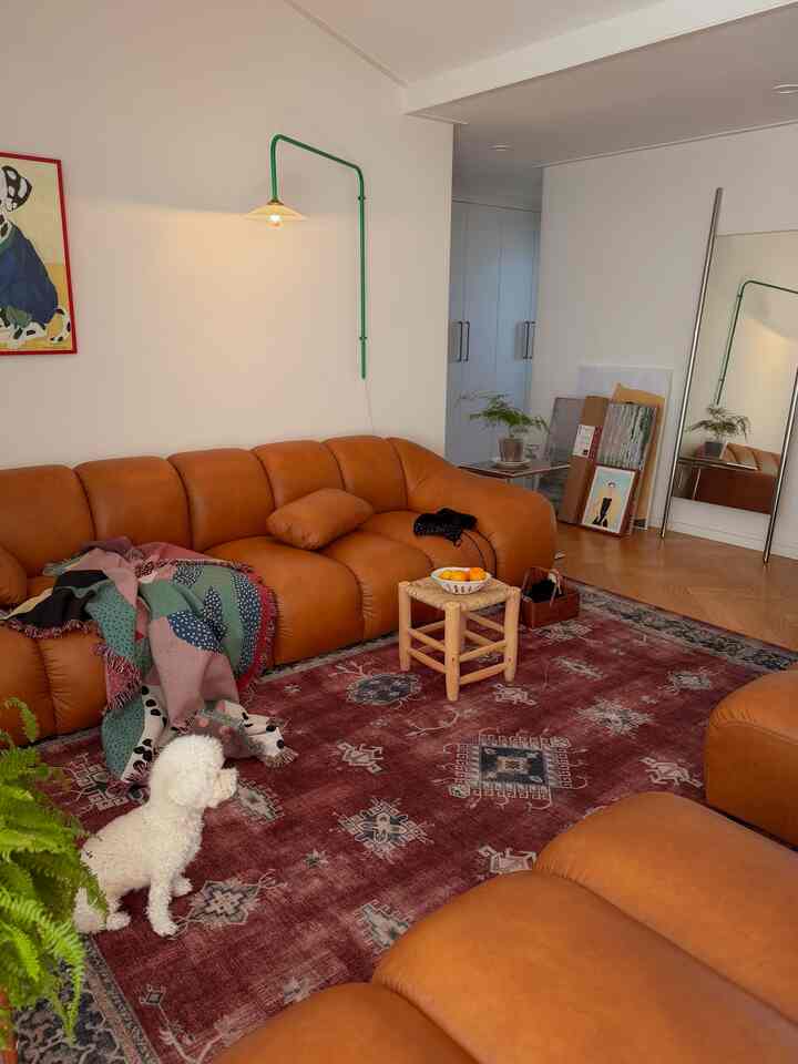 ナチュラルカラーの温かみのあるリビング、広いセクショナルレザーソファとパターンラグ、小さな丸いラタンのスツール、壁掛けランプが配置された空間