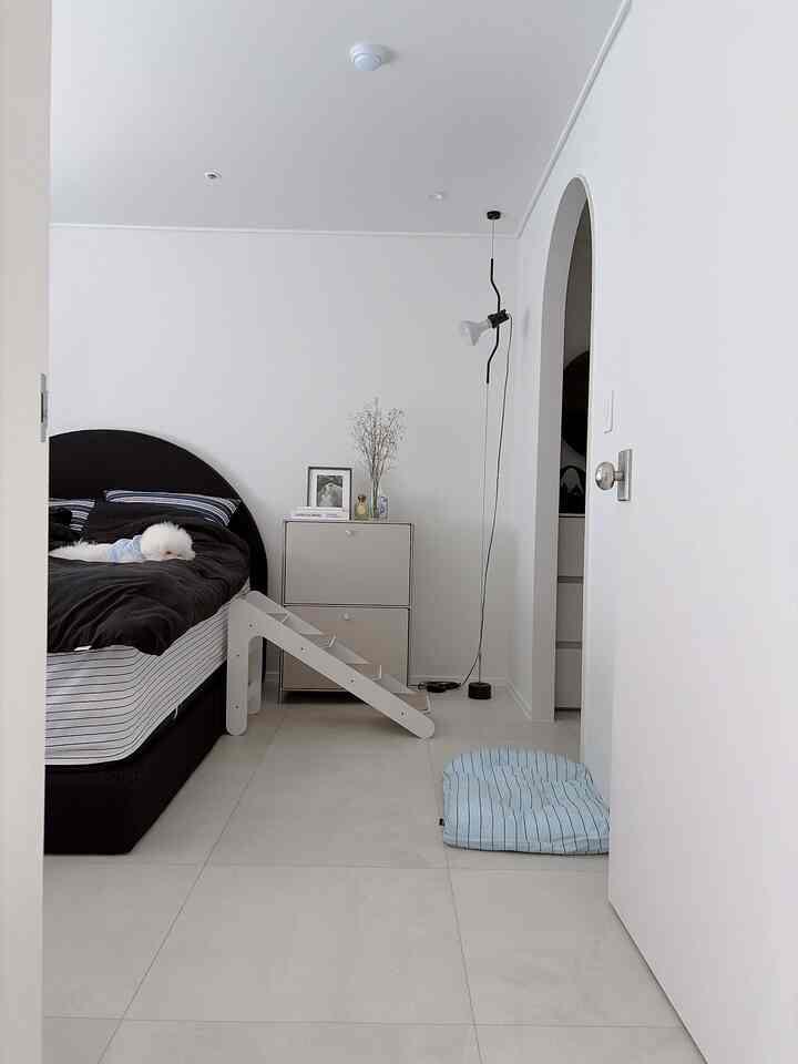 白い壁と黒いベッドフレームが調和したシンプルな寝室で、ペット用ステップとナイトテーブルが配置され、小型犬とともに使われる清潔感のある空間