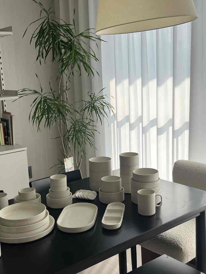 白いカーテンが背景のダイニングルーム、黒いダイニングテーブルの上に整然と並べられた白いセラミック食器が置かれたモダンな空間